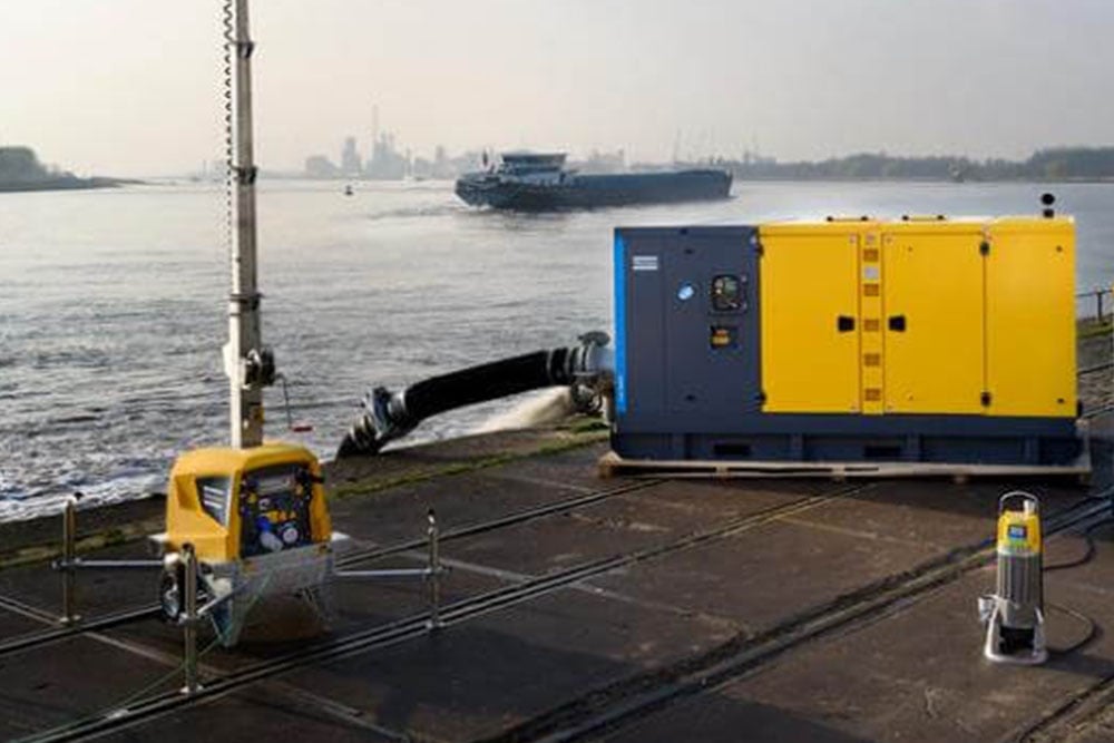 A large yellow and blue industrial pump system set up on a concrete platform beside a river, with hoses extending into the water. Smaller pump equipment is also visible nearby, and a cargo ship is seen in the background.