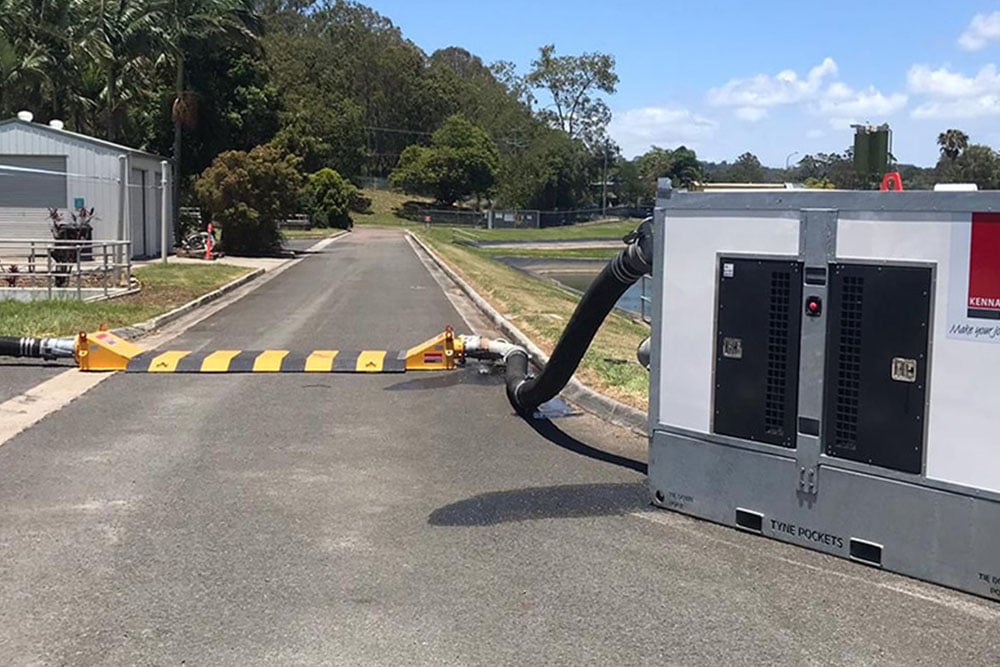 A large white and grey pump unit with the Kennards Hire logo, positioned beside a paved road. A black hose extends from the unit, running over a yellow and black cable ramp across the road, in a semi-rural setting with buildings and trees in the background.