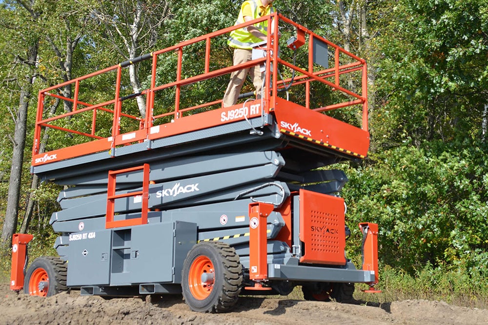 Scissor lift with a Kennards hire team member standing on the elevated platform in an outdoor, wooded area