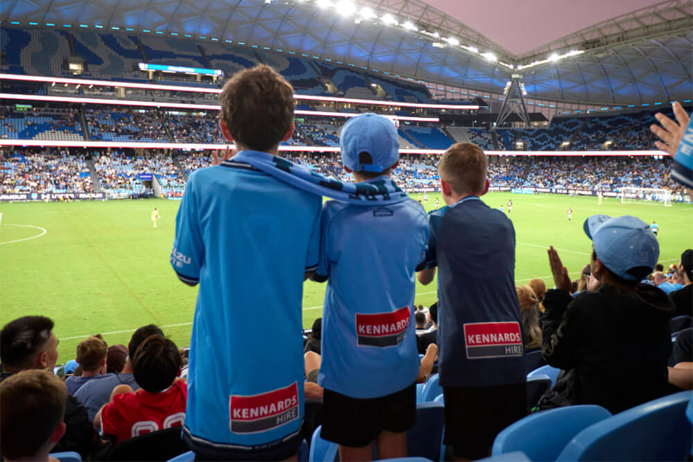 Three young Sydney FC fans standing and looking at the game from the stands
